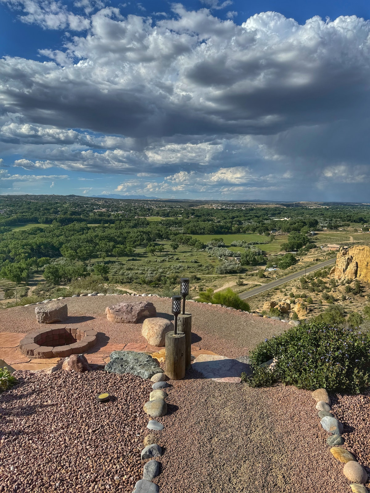 A scenic outdoor area features a fire pit surrounded by rocks, with views extending over lush green fields and distant hills. A pathway leads from the fire pit, and dramatic clouds hover above, reflecting the serene atmosphere of the landscape.