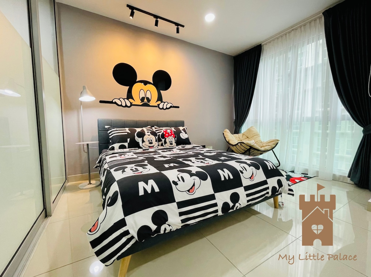 A sleeping area features a spacious bed adorned with a playful Mickey Mouse-themed duvet cover. Neutral walls are complemented by a decorative mural of Mickey Mouse. A floor lamp provides additional lighting, while large windows are dressed with dark curtains, allowing natural light to filter through.