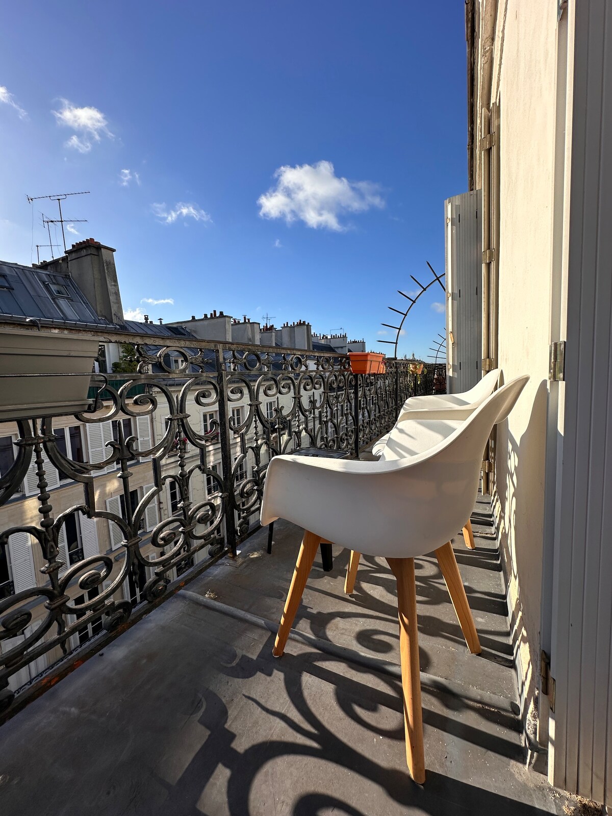 A sunlit balcony is shown with a single white chair featuring wooden legs. Ornate black wrought iron railings frame the view, with a clear blue sky and scattered clouds in the background, creating an inviting outdoor space.