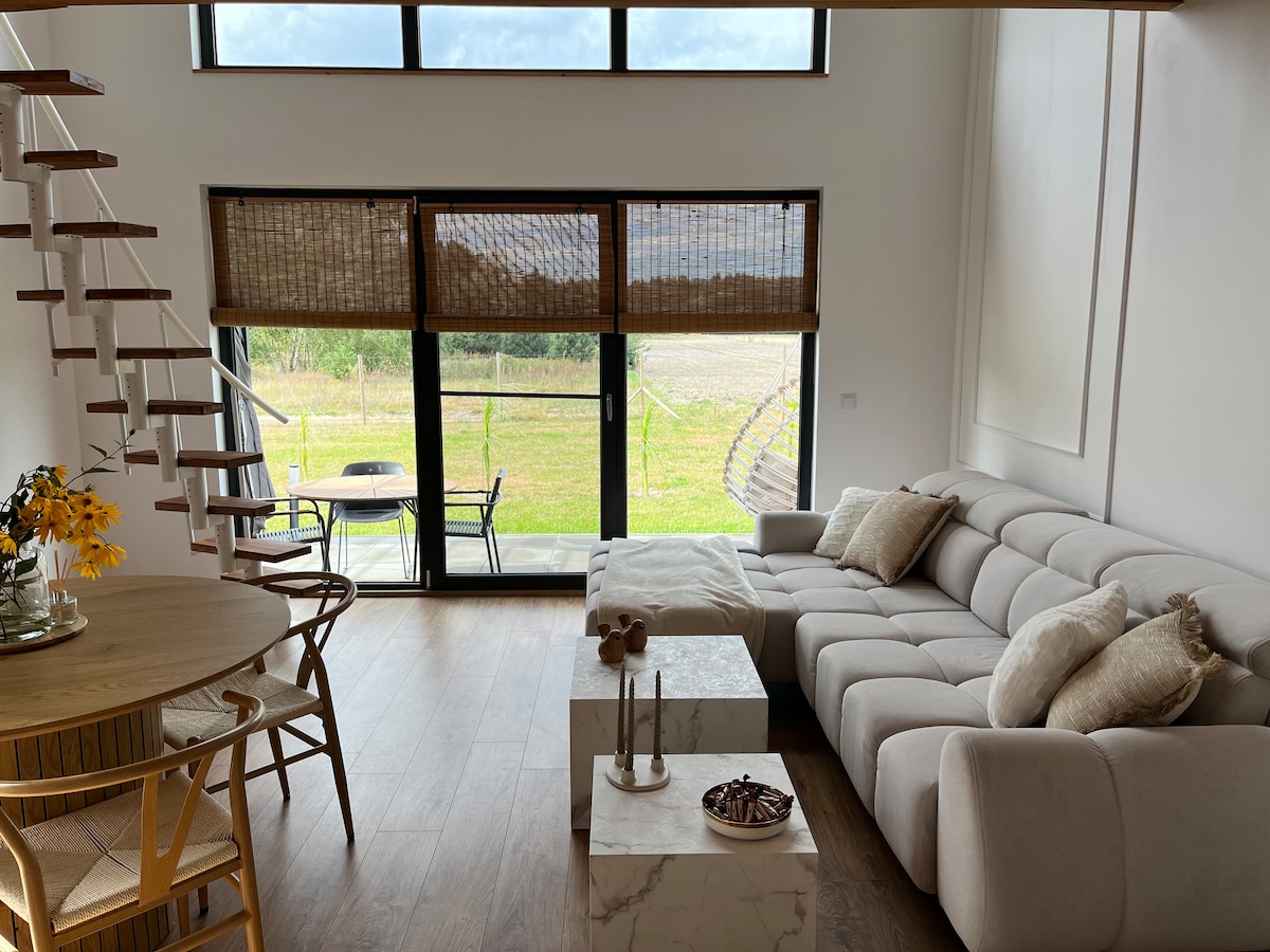 A modern living area is showcased, featuring a light neutral sofa, a marble coffee table, and a round dining table with rattan chairs. Large windows let in natural light, offering views of the outdoors, while warm blinds provide desired privacy.