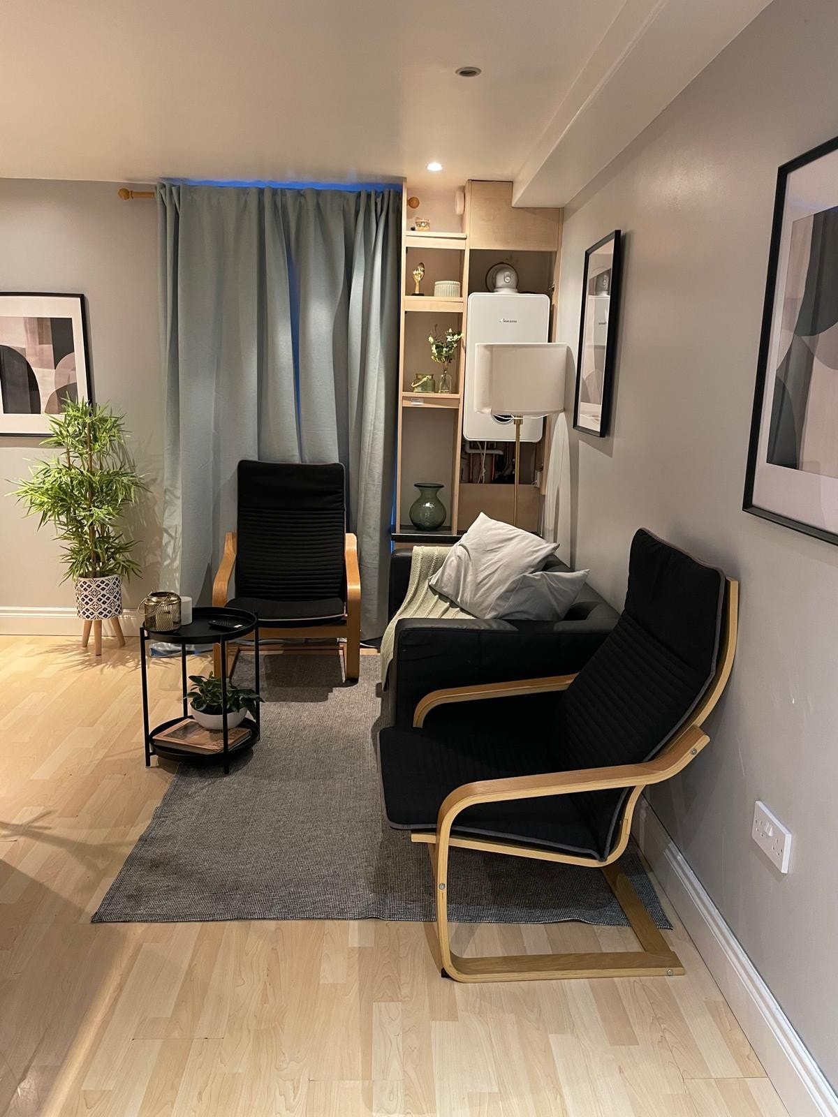 A cozy sitting area is illustrated with two minimalist chairs positioned beside a small, round side table. A plant adds a touch of greenery, while light curtains frame a window, creating a serene environment. Neutral artwork is displayed on the walls, enhancing the modern aesthetic.