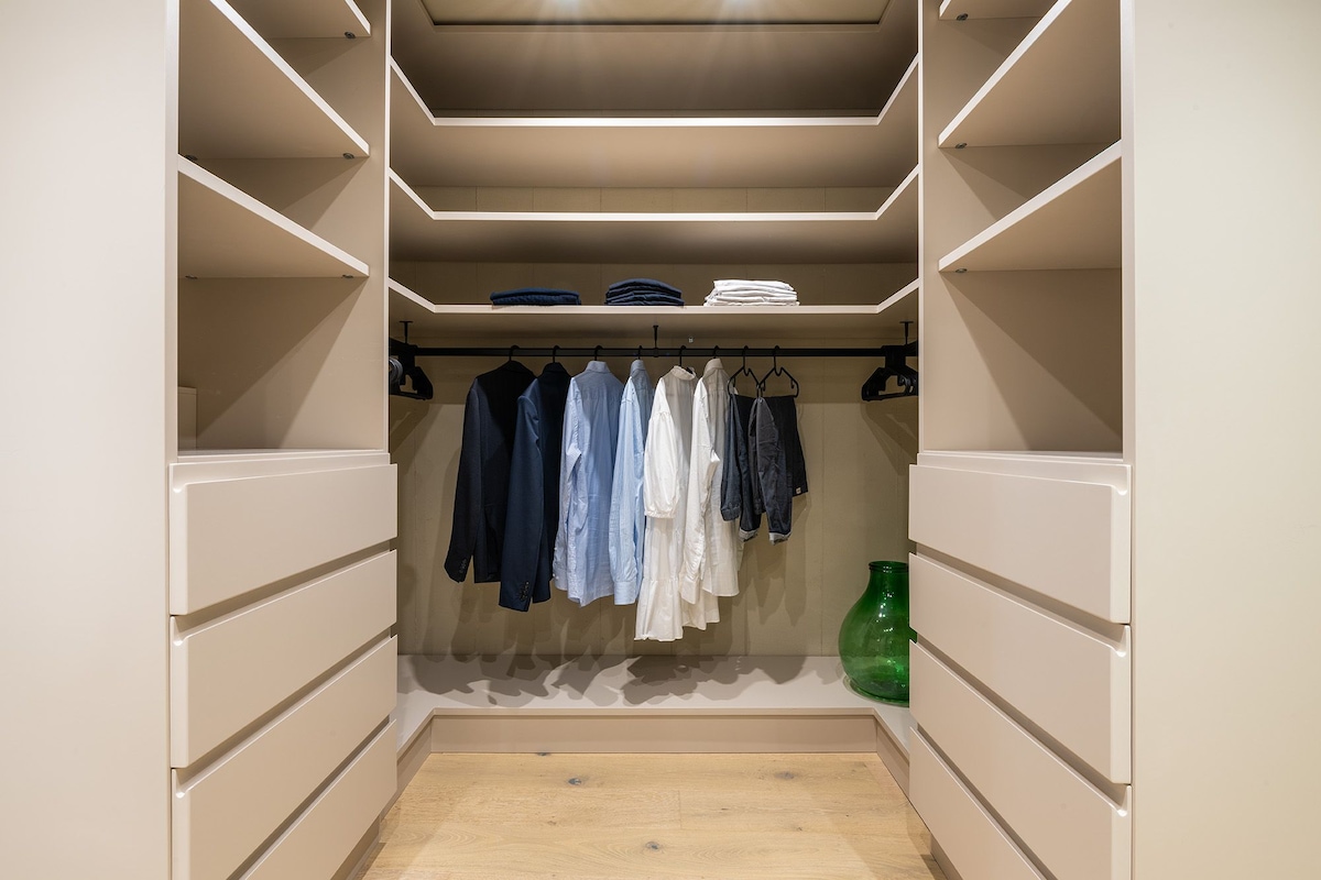 A spacious walk-in closet is presented with organized shelves and hanging space. Neatly arranged clothing items in various colors are visible, alongside several drawers below. A green decorative item adds a subtle touch to the neutral-toned interior.