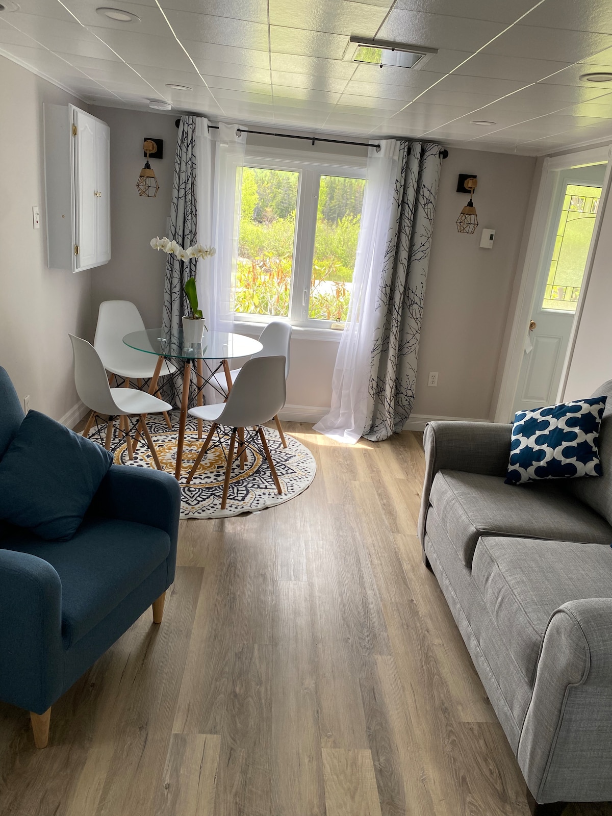 A comfortable seating area is featured, with a gray sofa and a blue armchair situated around a round glass table. Natural light filters through the large window, complemented by sheer curtains, while a patterned rug enhances the warmth of the space.