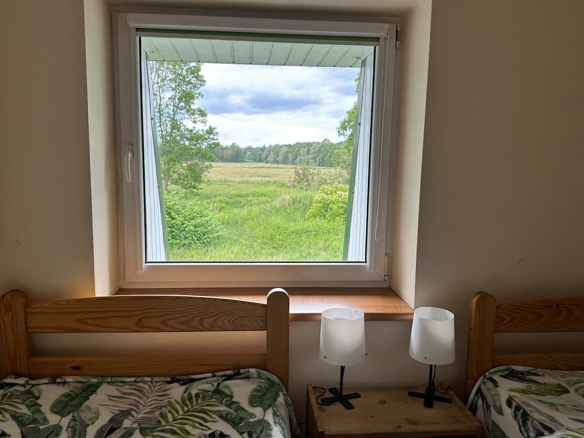A window with a clear view of a green landscape showcases fields and trees. Two bedside tables with lamps are positioned beside two twin beds, featuring floral patterned bedding. Natural light floods the room, enhancing the connection to the outdoors.