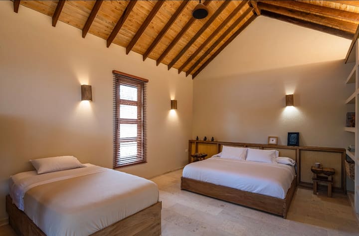 Habitación rústica de estilo caribeño con detalles naturales, muebles de madera y tonos cálidos. Incluye una cama tamaño king y cuatro camas individuales, perfectas para familias o grupos que buscan comodidad y tranquilidad en un ambiente tropical acogedor.