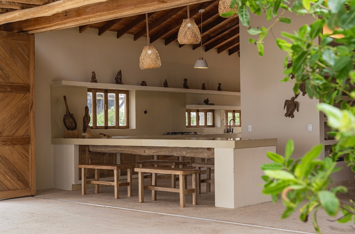 An open kitchen space features a large wooden dining table with benches, surrounded by warm, natural light from multiple windows. The area is adorned with woven pendant lights and rustic decor, creating a welcoming environment for gatherings.