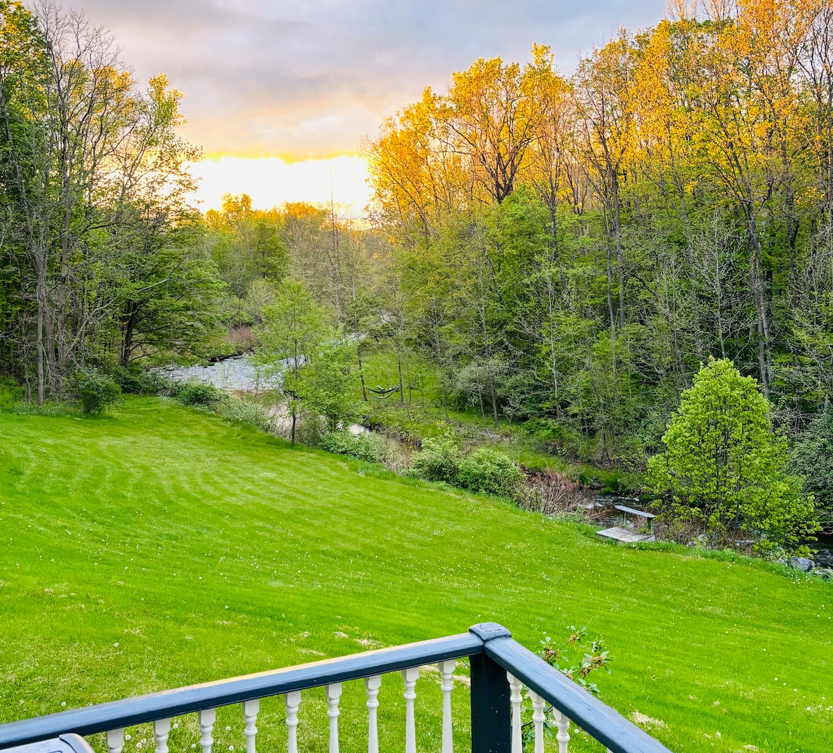 A serene view of a lush green lawn extends towards a gently flowing creek, bordered by trees in various shades of green. The setting sun casts warm light across the landscape, highlighting the natural beauty of the surrounding woods.