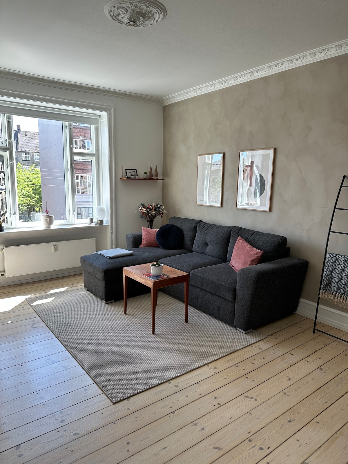 A comfortable living area is presented with a dark sectional sofa and a small wooden coffee table. Two framed artworks adorn the light-colored walls. A window allows natural light to fill the space, complemented by a rug on the wooden floor.