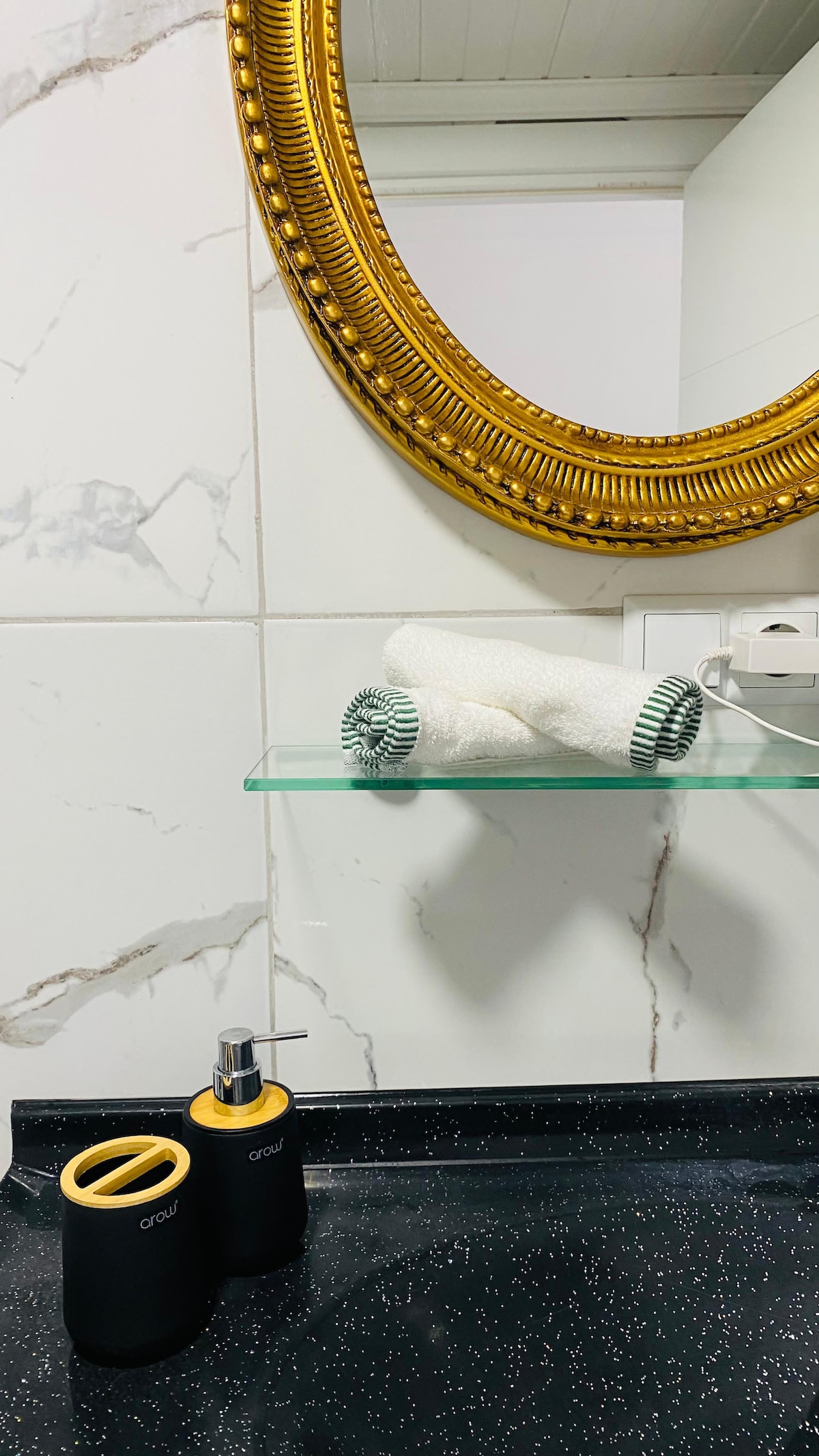 A modern bathroom features a circular mirror with a textured gold frame above a glass shelf holding rolled white towels. Two minimalist black and gold containers are positioned on a dark countertop, detailing a sleek and organized space.