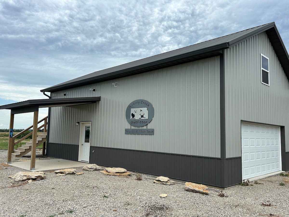 A modern two-story building with a sleek metal exterior is displayed. A large garage door is visible on one side, and a welcoming entrance with steps is identifiable. The surrounding area features a gravel surface and distant greenery.