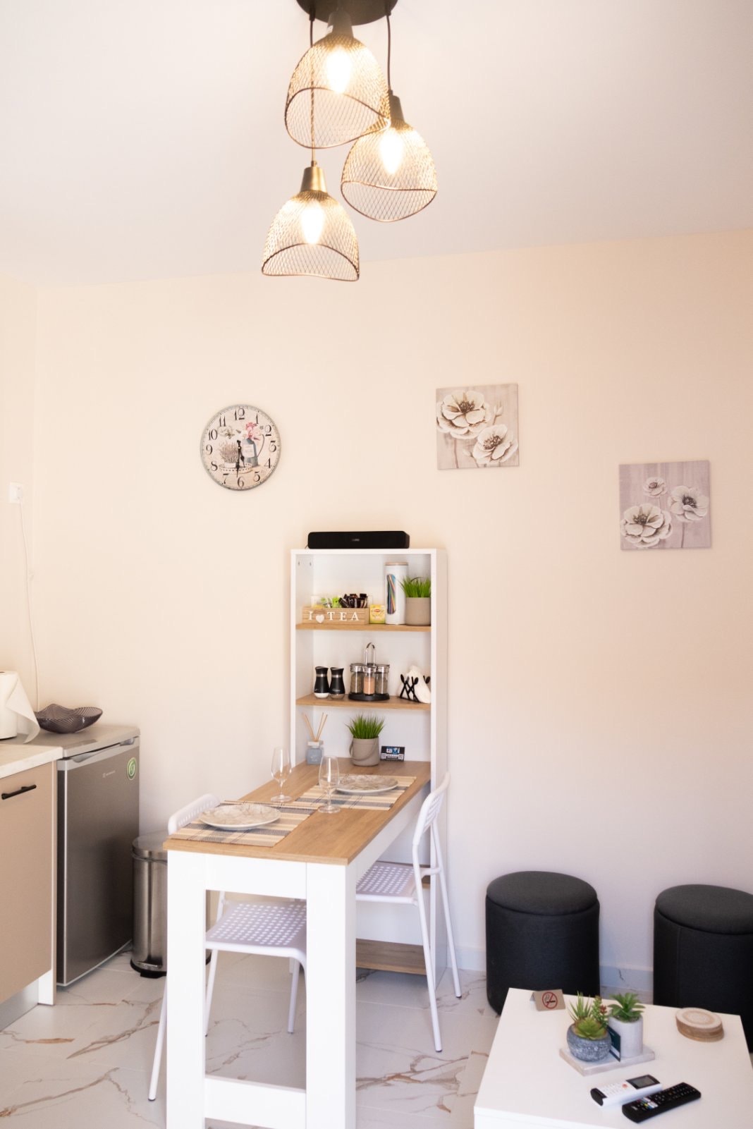 A modern kitchen area features a light wood dining table surrounded by white chairs. Wall art adds subtle decor above a compact shelf, which holds kitchen essentials. Soft lighting is provided by three pendant lamps hanging from the ceiling, enhancing the bright and functional space.