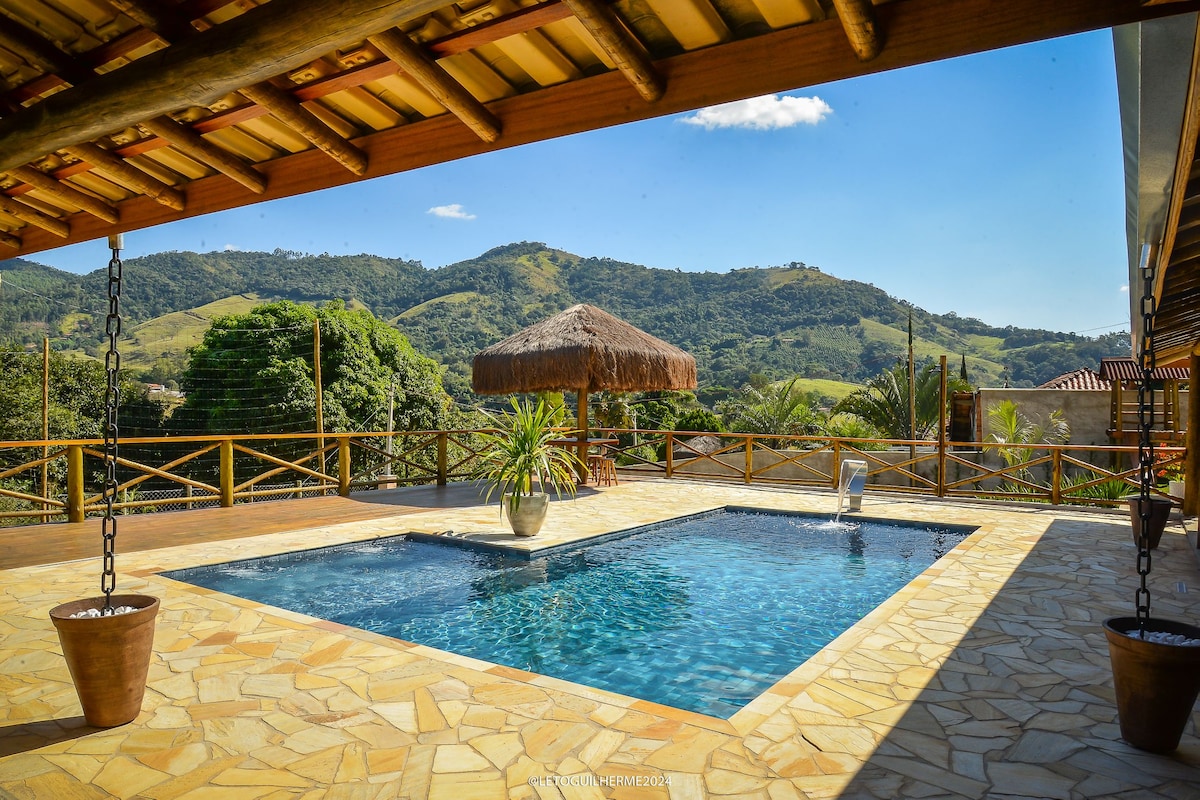 A spacious outdoor area showcases a tranquil pool surrounded by stone tiling. Lush green mountains roll in the background under a clear blue sky, while a thatched-roof gazebo adds to the serene atmosphere. Sunlight reflects on the water, enhancing the inviting setting.