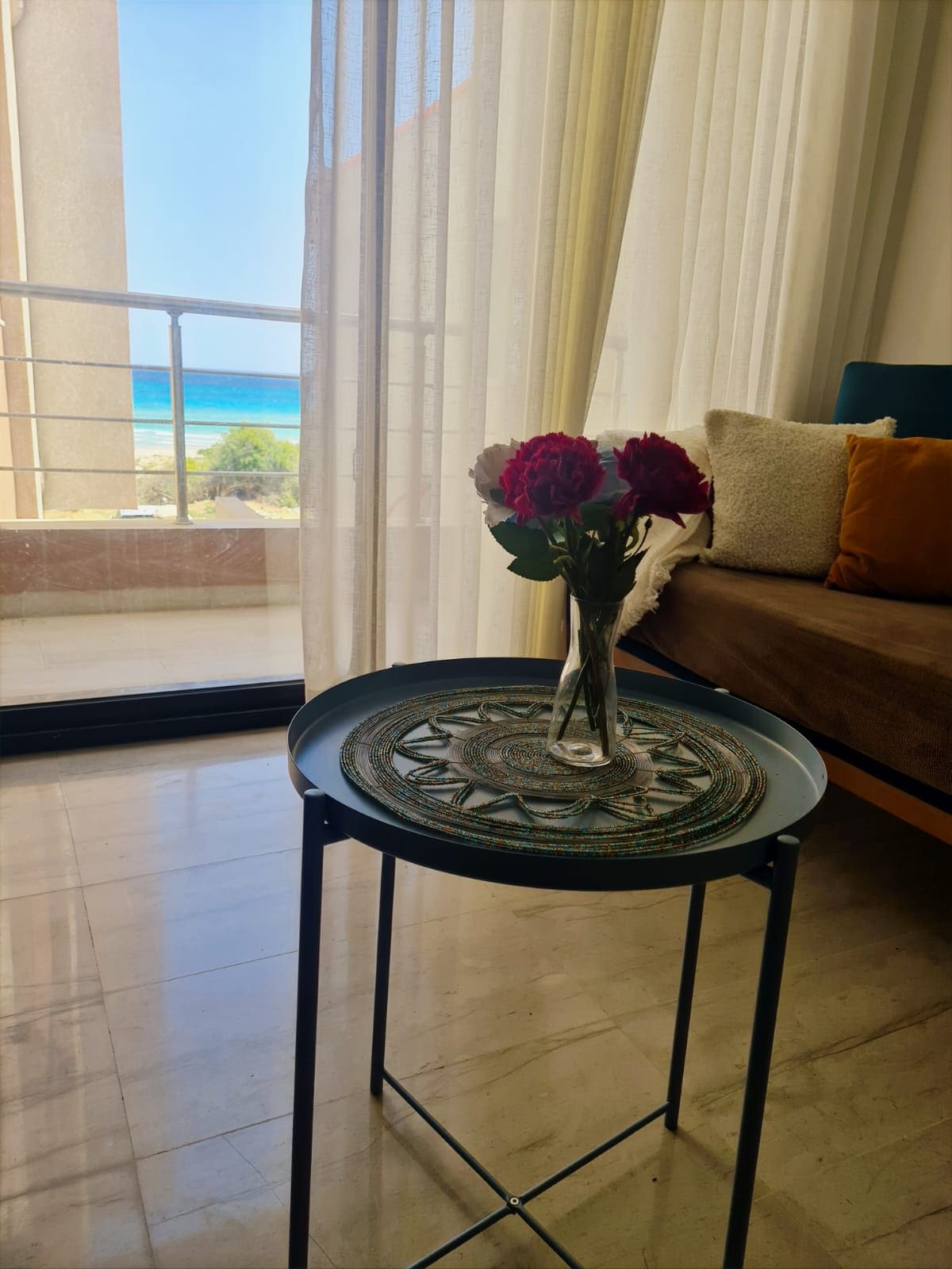 A small, round table is set with a decorative tray and a vase holding fresh flowers. A comfortable sofa with accent pillows is positioned nearby, and sheer curtains frame a view of the sea through glass doors.