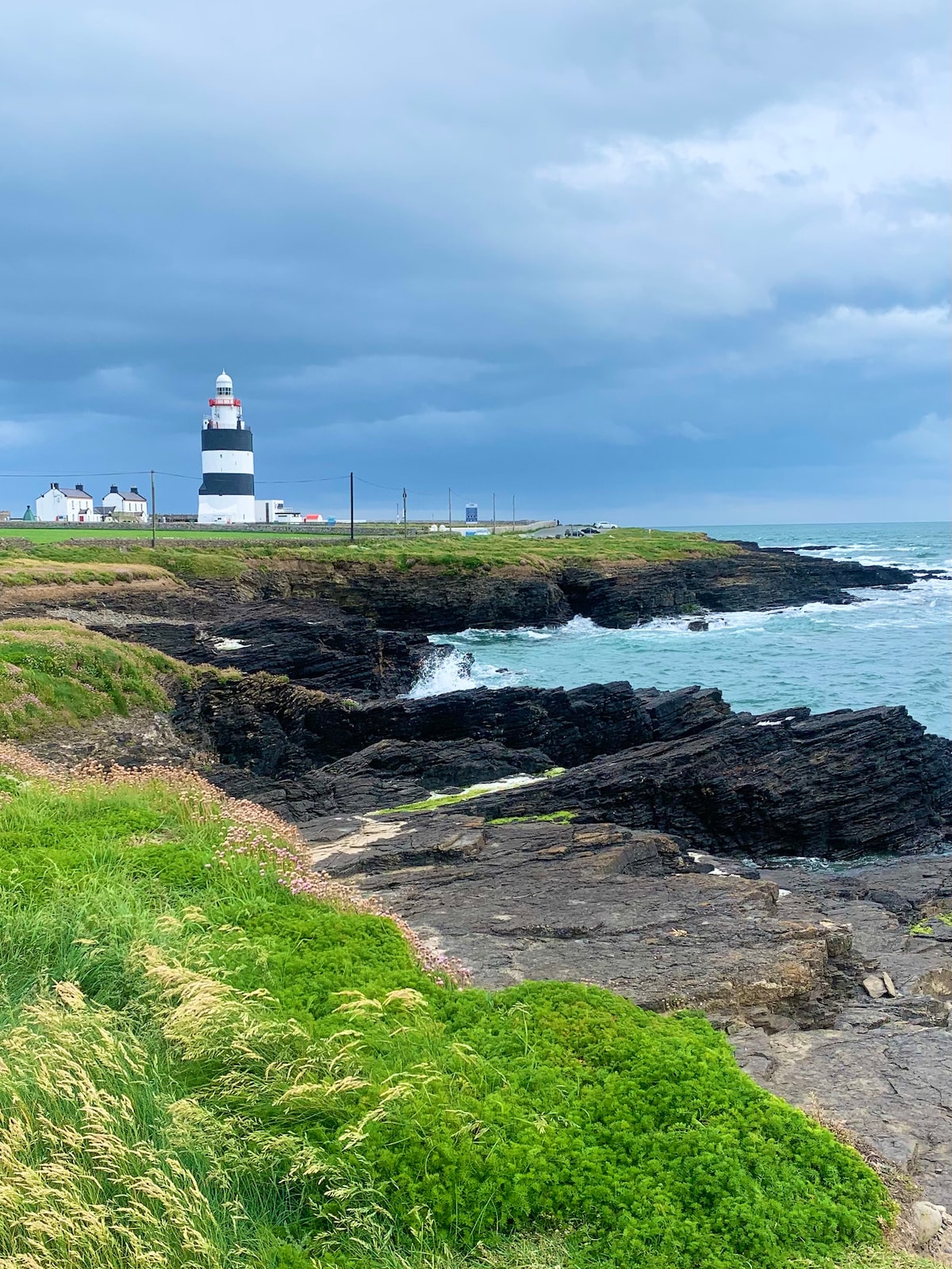 A striking coastal scene features a traditional lighthouse with black and white stripes, standing along the rugged shoreline. The rocky outcrops extend into the churning sea, while lush greenery frames the image. The sky is overcast, hinting at a breezy atmosphere.