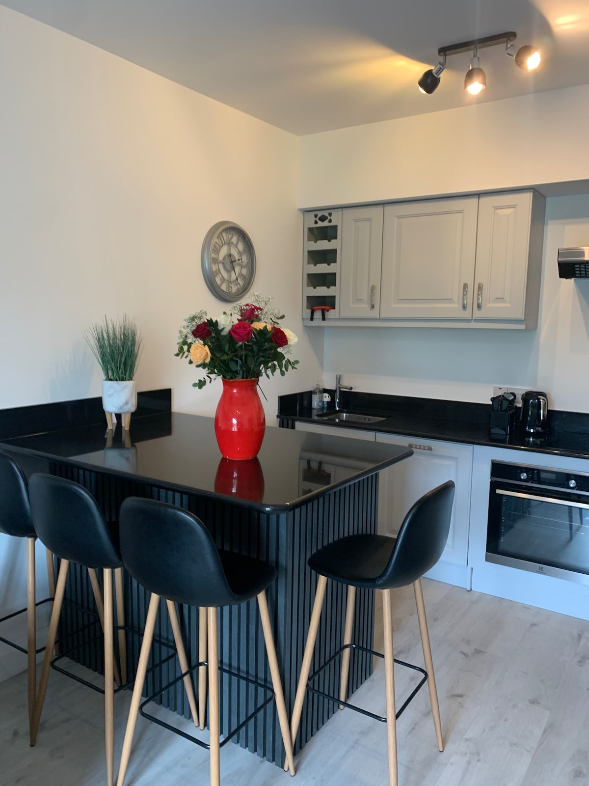 A modern kitchen is featured with a sleek black countertop and four minimalist bar stools. Decorative flowers are arranged in a vibrant red vase, complementing a stylish clock on the wall. Kitchen appliances, including a coffee maker and oven, are neatly positioned for convenience.
