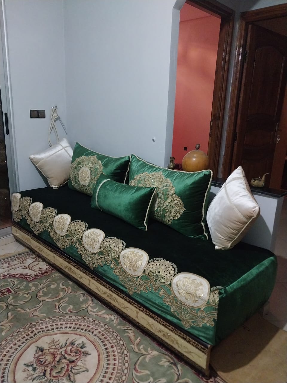 A stylish seating area features a long, plush sofa adorned with intricate golden embroidery. The sofa is complemented by decorative cushions, and a colorful area rug lies beneath it. Soft lighting creates a cozy atmosphere in the room.