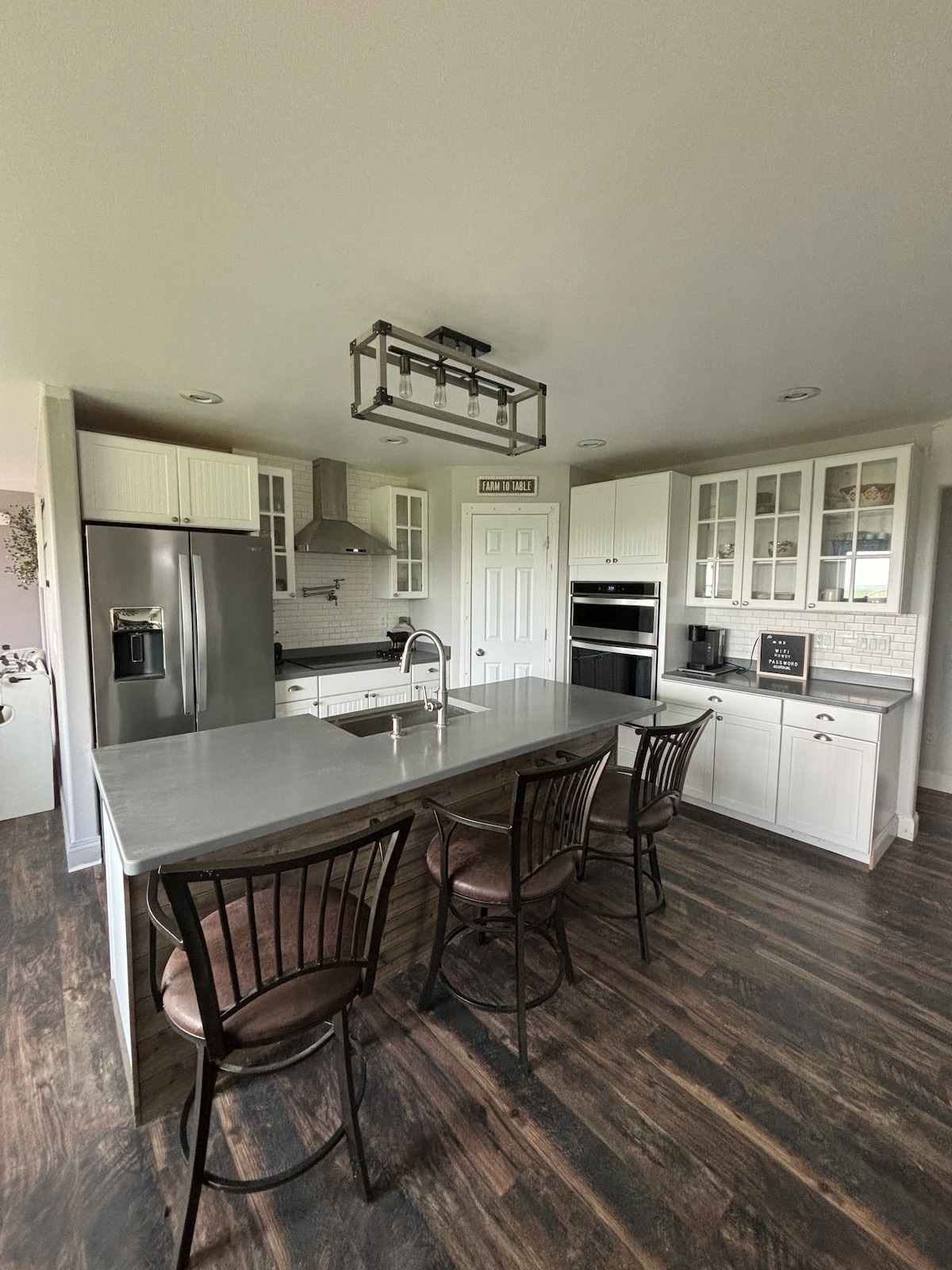A modern kitchen features sleek white cabinetry and stainless-steel appliances. An island with four high-backed chairs provides ample seating. The space is well-lit, with a stainless-steel hood above the cooktop and a cozy ambiance created by soft under-cabinet lighting.