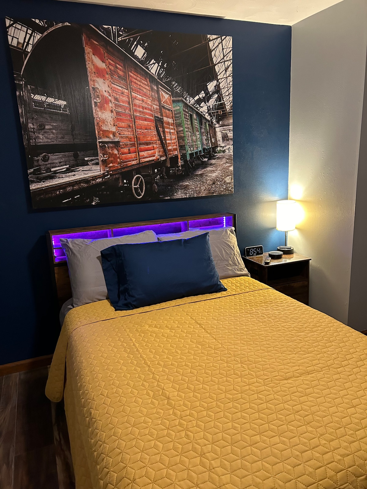 A cozy bedroom features a full-sized bed dressed in a yellow quilt, complemented by a blue accent pillow. A stylish nightstand holds a lamp, and a striking wall photograph of colorful train cars adds character to the room. Soft lighting creates a warm ambiance.