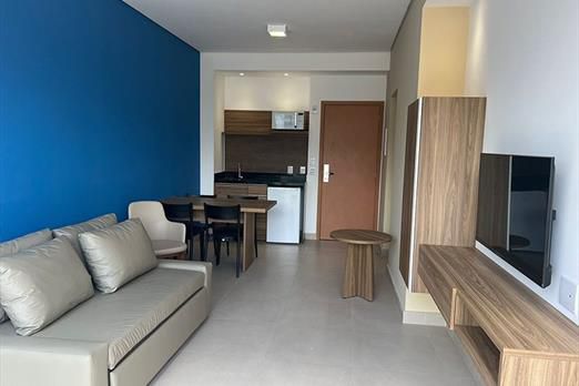 A modern living area features a comfortable sofa and a small wooden table. A dining set for four is positioned adjacent to a compact kitchen area with essential appliances. A flat-screen TV is mounted on the wall, enhancing the overall design.