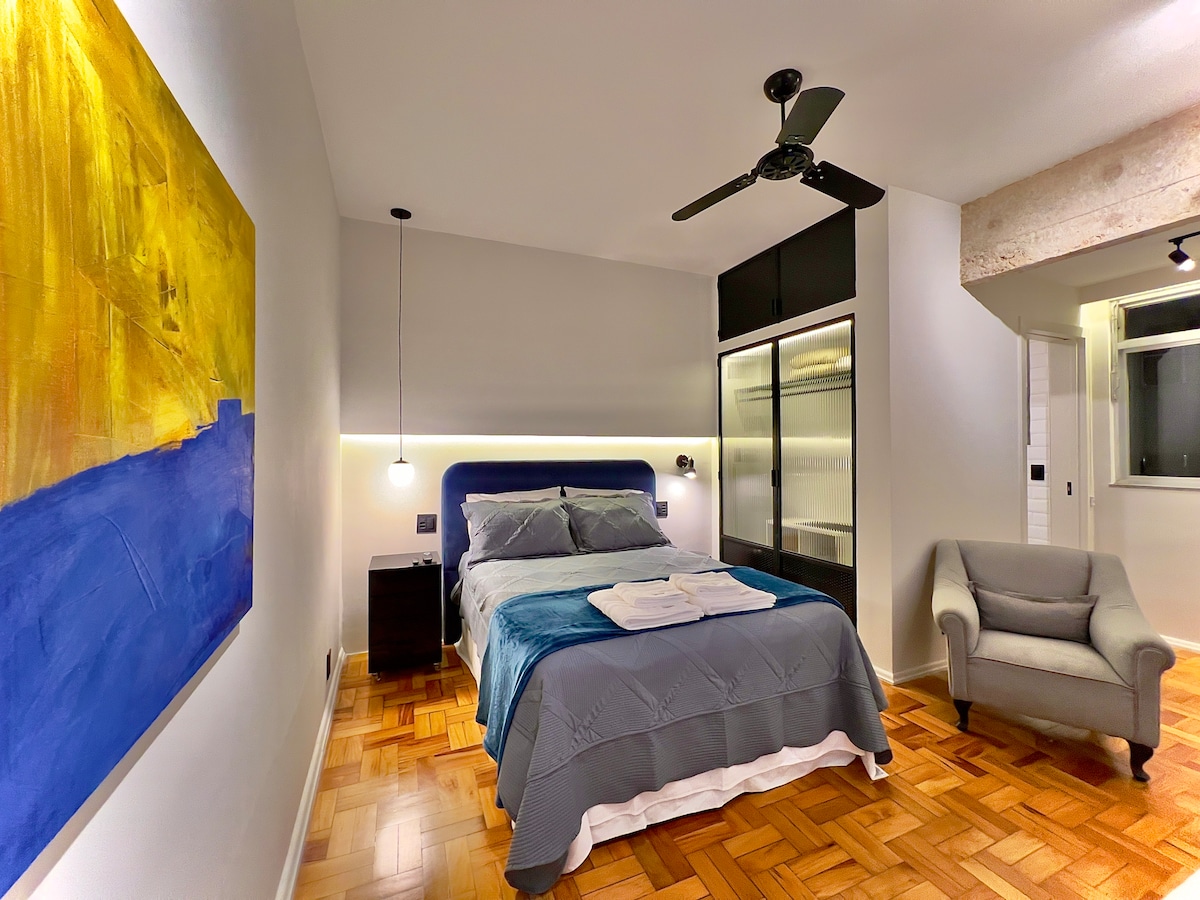 A comfortable sleeping area is presented, featuring a spacious bed with blue and gray bedding. A soft armchair sits in the corner, and a colorful wall painting adds a pop of color. A ceiling fan hangs above, providing additional airflow.
