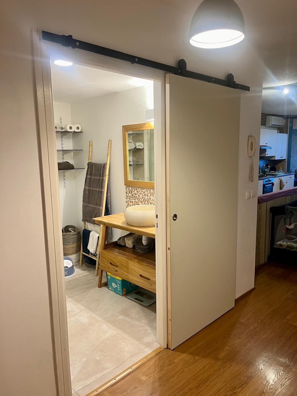 A sliding barn door leads into a bathroom area featuring wooden shelving with neatly arranged towels and toiletries. A decorative mirror above a wooden vanity reflects the light from a ceiling fixture. Pale tile flooring adds to the clean and organized space.