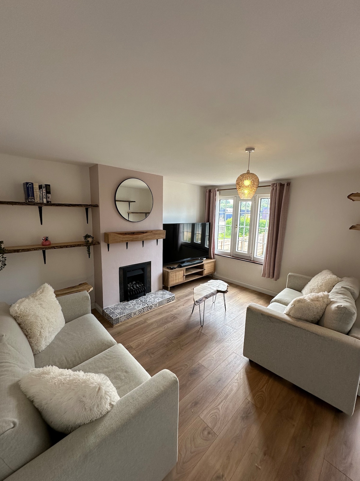 A cozy living room features two light-colored sofas adorned with soft cushions. A round mirror is mounted above a modern fireplace, while a television is positioned on a contemporary unit. Natural light filters through the windows, accompanied by neutral-toned curtains.