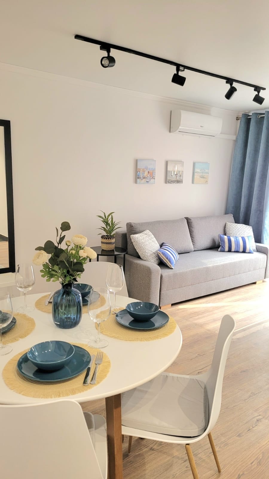 A stylish living area features a gray sofa, accented by decorative cushions. A round dining table is set with blue dishware and a floral centerpiece, surrounded by white chairs. Wall art adds a touch of character, while a large mirror enhances the sense of space.