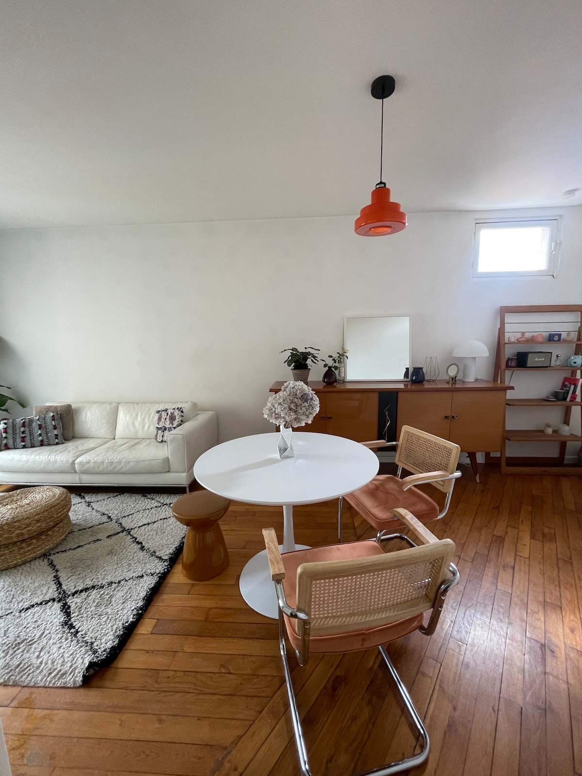 A light-filled living area is showcased with a circular white dining table set for two. Nearby, a soft cream-colored couch offers seating. A wooden sideboard, a wall mirror, and vibrant greenery are present, contributing to a relaxed atmosphere. Warm wooden flooring adds to the inviting feel.
