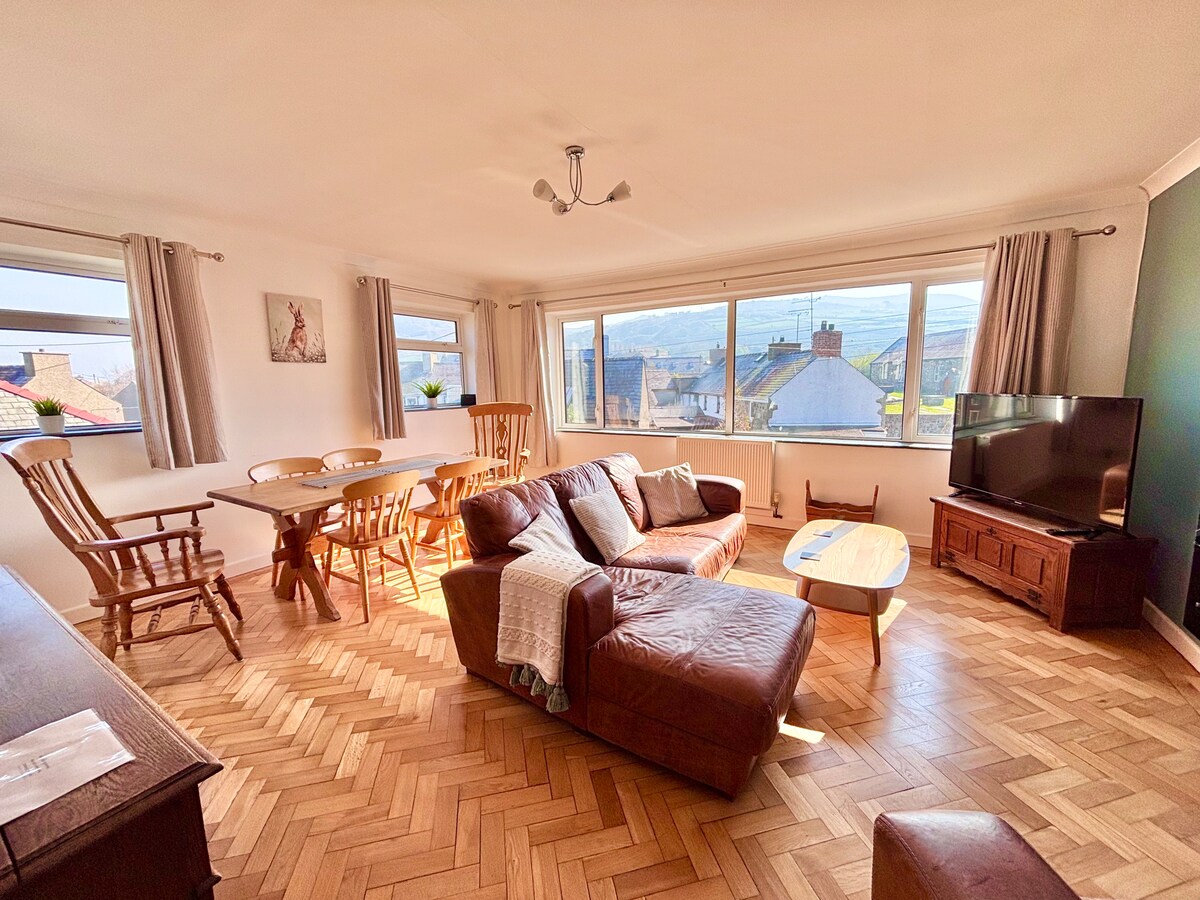 A bright living and dining area is presented with large windows allowing natural light to fill the space. A comfortable leather sofa is accompanied by a wooden coffee table. A dining table with wooden chairs is visible, creating a relaxed setting.