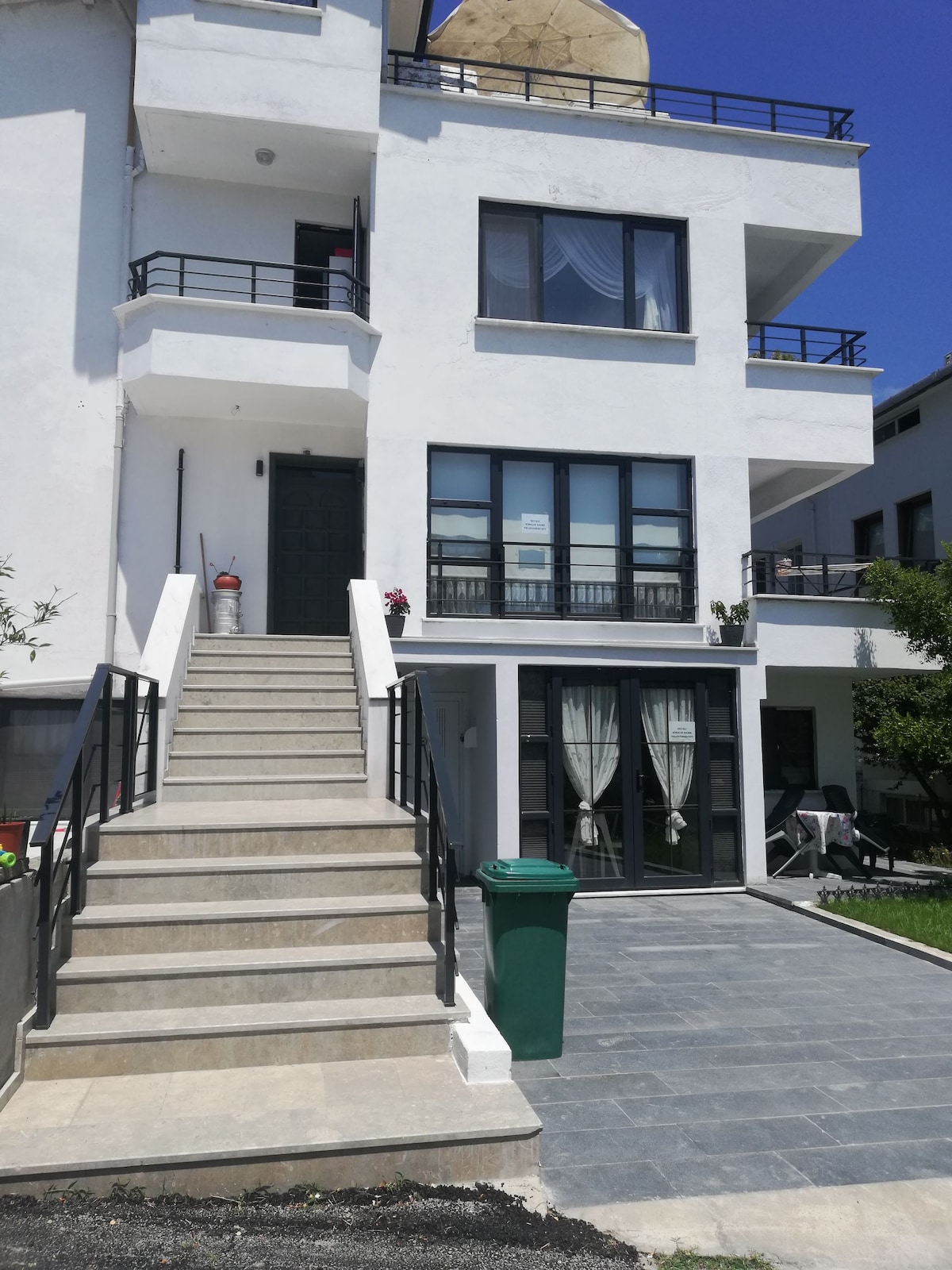 A modern three-story building features a spacious outdoor staircase leading to the entrance. Large glass windows allow natural light to fill the interior. Green landscaping accents the exterior, with a trash bin positioned at the base of the stairs.
