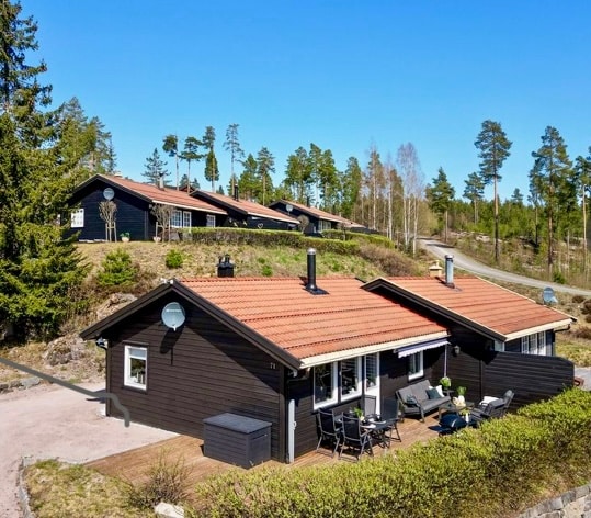 A cluster of charming black wooden cabins is surrounded by greenery and tall trees. Brightly colored rooftops contrast with the natural landscape. Each cabin features a private terrace furnished with outdoor seating, inviting relaxation in a tranquil outdoor setting.