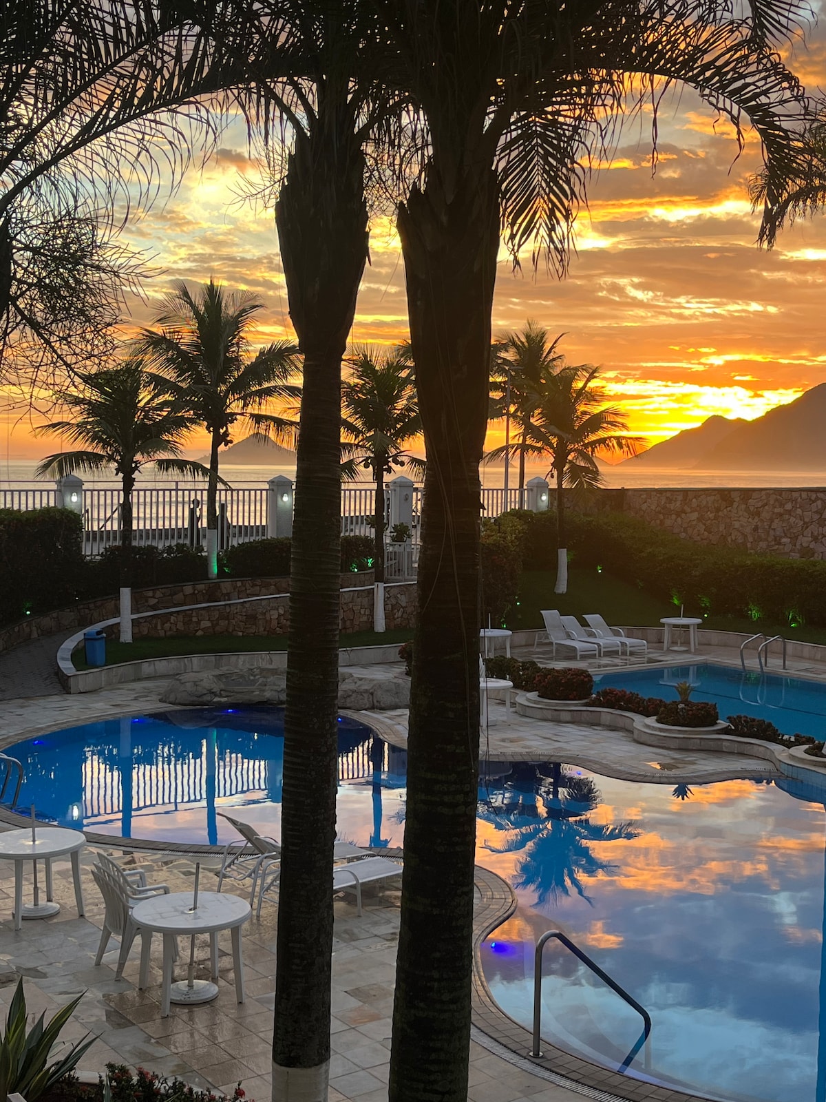 A serene pool area is framed by tall palm trees, reflecting vibrant hues of sunset in the water. Outdoor seating is arranged around the pool, and the ocean is visible in the background, enhancing the tranquil atmosphere of the space.