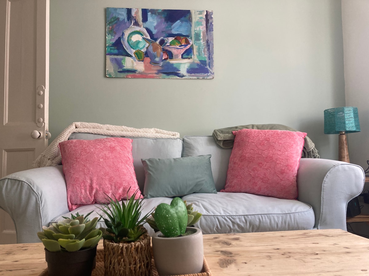 A comfortable couch is adorned with colorful throw pillows in soft pink and green shades. A small table holds an assortment of potted plants, adding a touch of greenery. A vibrant piece of art hangs on the wall, providing a pop of color to the light-hued room.