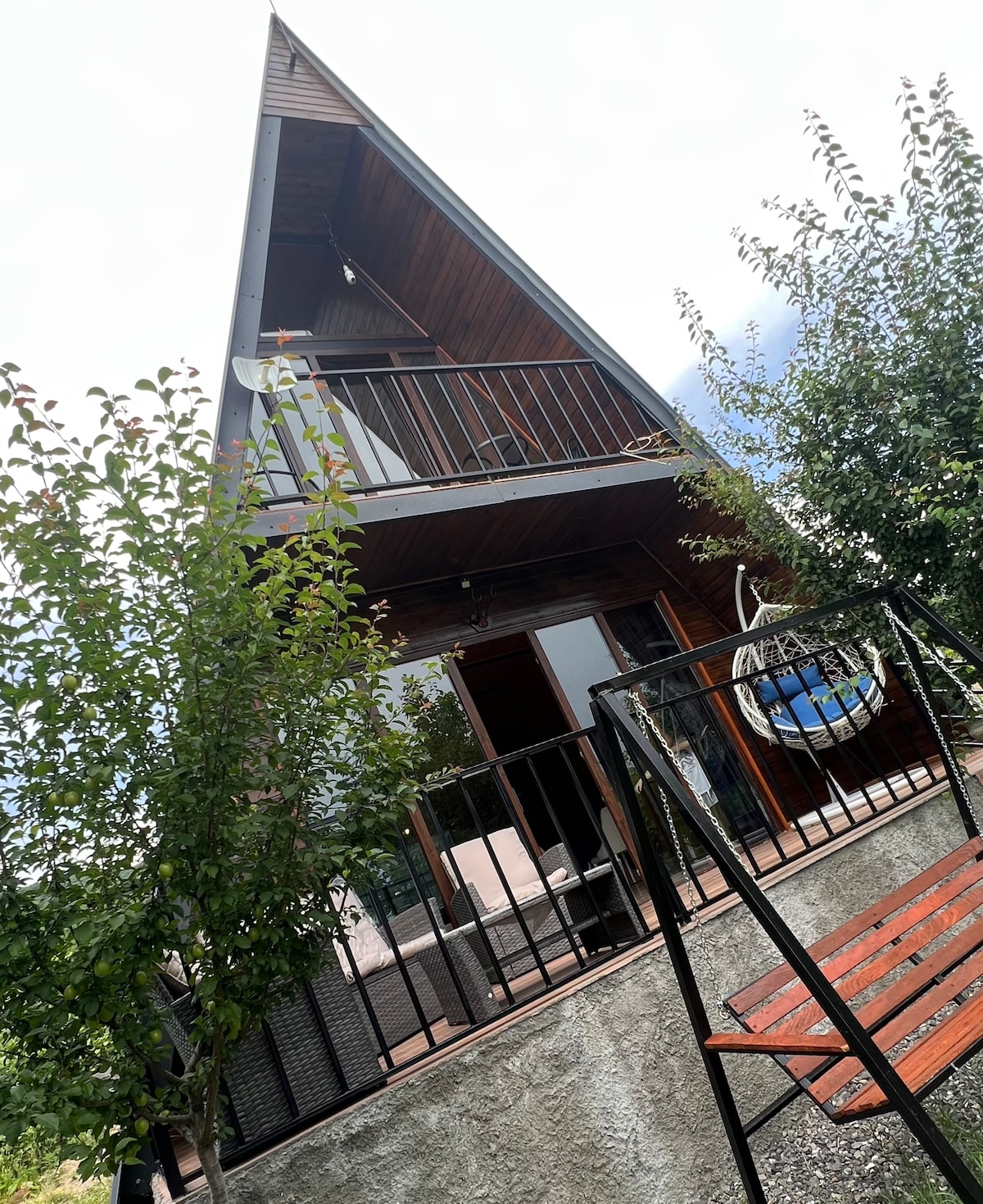 The A-frame cottage is surrounded by trees, featuring a stylish wooden exterior. A small balcony with a railing and outdoor seating provides a view of the surrounding greenery. The entrance is accessed by a short set of steps leading to sliding glass doors.