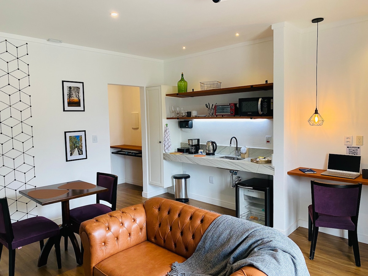 An integrated living space showcases a cozy seating area with a tan leather sofa and two small tables. A compact kitchen features essential appliances, including a microwave and coffee maker, set against a clean white background. Subtle lighting enhances the inviting ambiance.