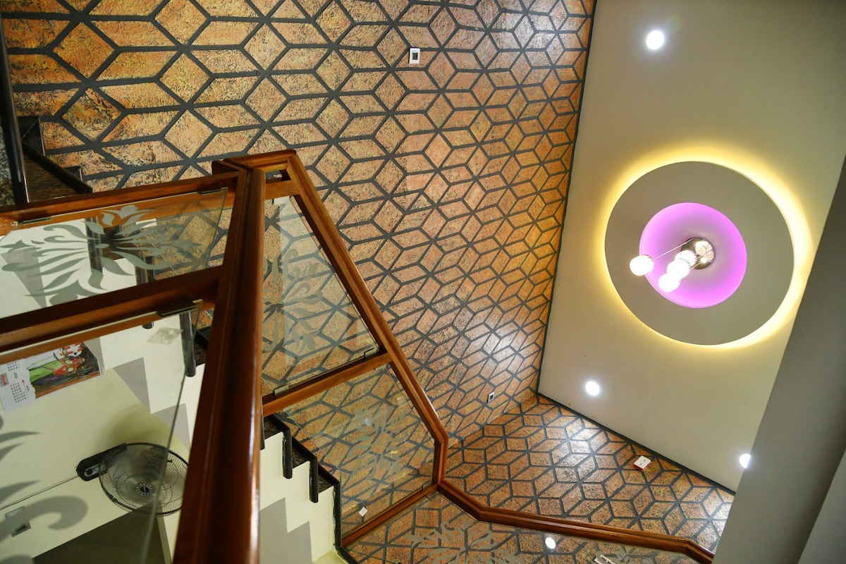 A stylish staircase is surrounded by a geometric patterned floor, leading to an upper level. A circular light fixture with a purple accent illuminates the ceiling, enhancing the modern design. The railing is crafted from wood and glass, providing a clear view of the space.
