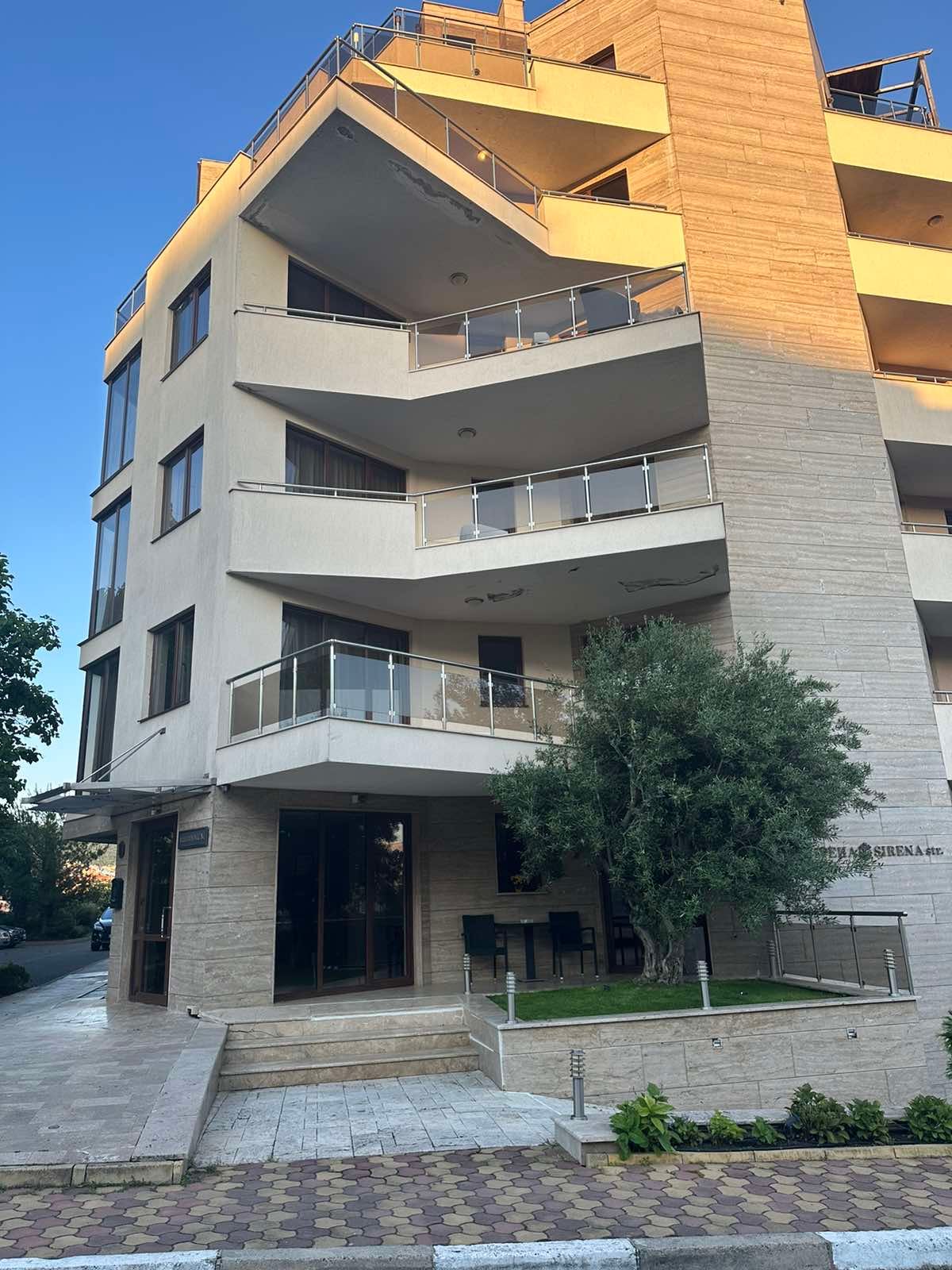 The modern building features multiple balconies with glass railings, promoting outdoor enjoyment. A stone facade complements the landscaping, which includes a small garden area at the entrance. Large windows enhance natural light, creating an inviting atmosphere.