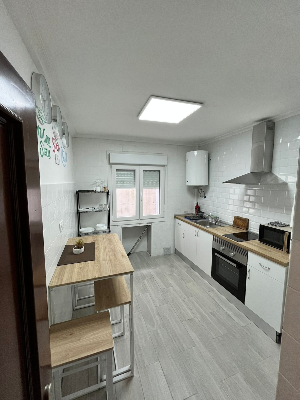 A modern kitchen area is presented, featuring light-colored cabinetry and sleek appliances. A small wooden table with two stools is positioned near a window, allowing for natural light. The space includes a microwave and an oven built into the cabinetry, providing functionality.