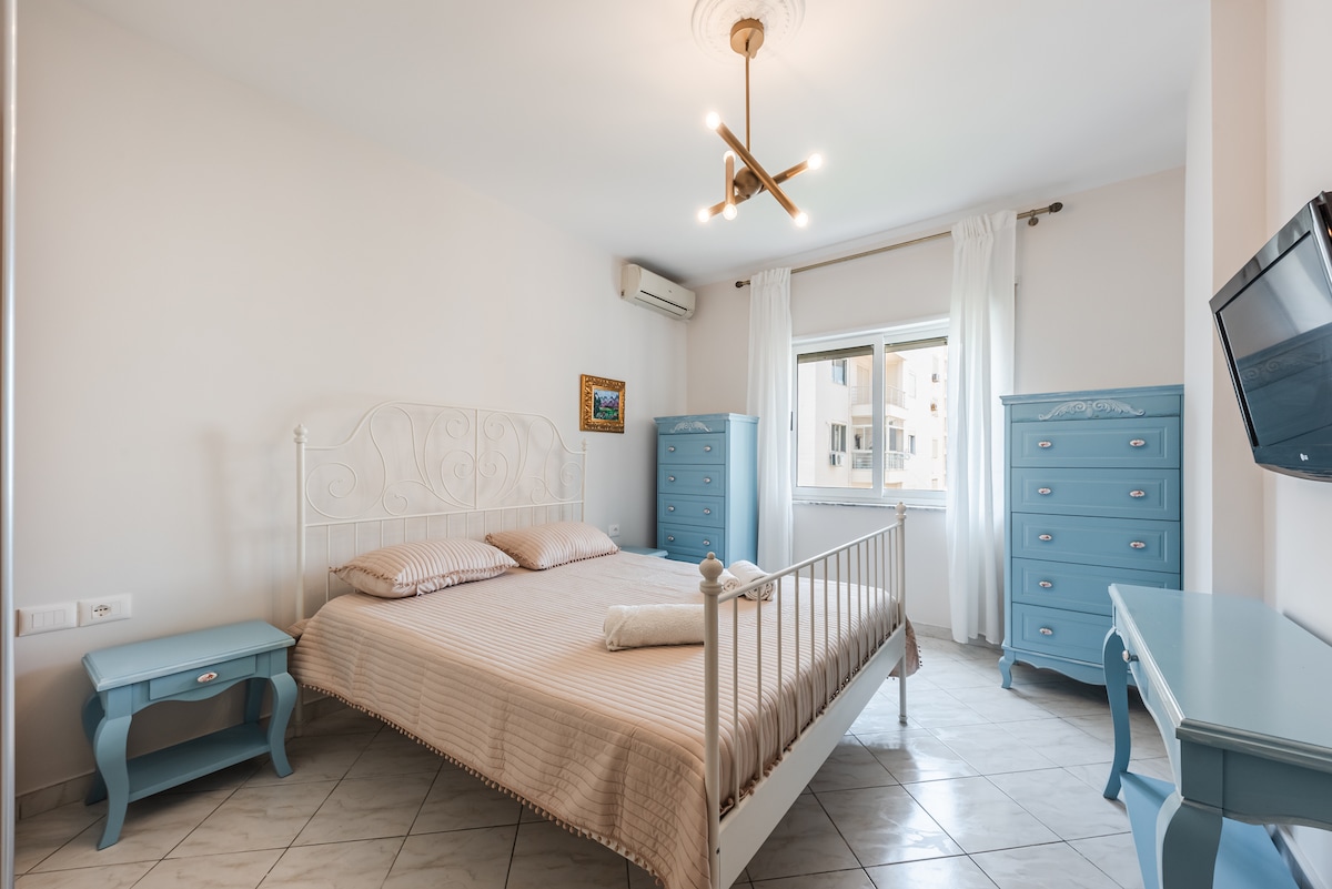 A cozy bedroom is featured, furnished with a spacious bed covered in a light-colored bedding set. Two stylish blue dressers are positioned on either side, and a flat-screen TV is mounted on the wall. Natural light fills the room through a window with sheer curtains.