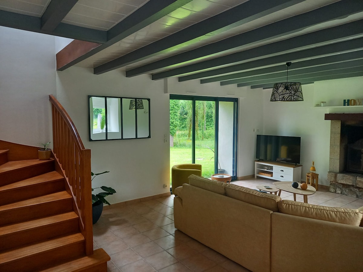The spacious living area features wooden beams and large glass doors overlooking the garden. A neutral-colored sofa and a TV are positioned nearby. A staircase leads to the upper floor, accompanied by a decorative plant and a mirror. Natural light fills the space.
