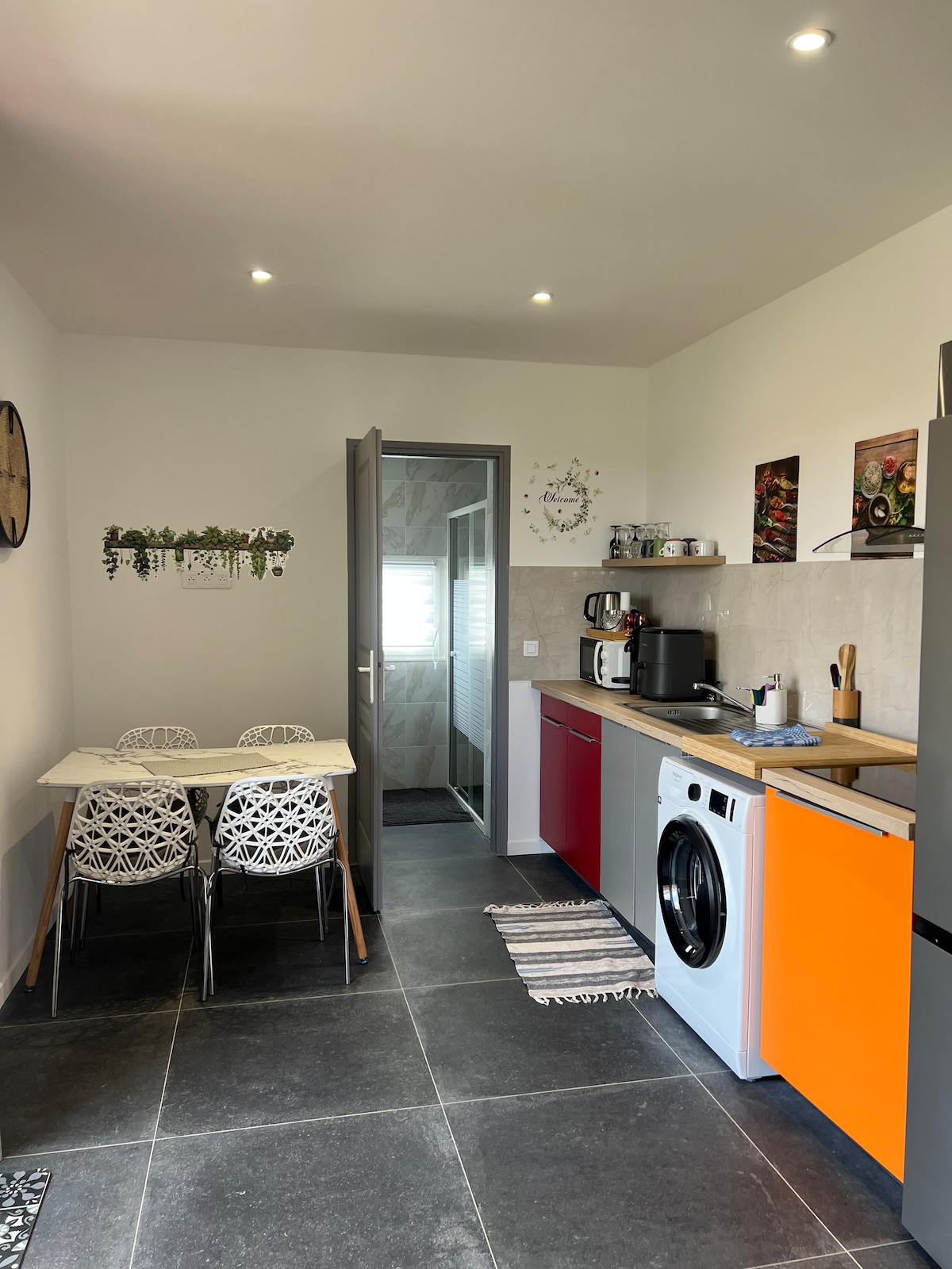 A modern kitchen features a compact dining table with two stylish chairs. An assortment of appliances is visible, including a washing machine and a coffee maker. The kitchen is illuminated by recessed lighting, and stylish wall decorations add personality to the space.