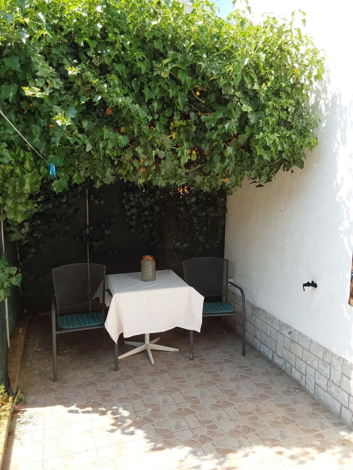 A shaded outdoor seating area is depicted, featuring a round table with a white tablecloth at its center. Two chairs are arranged around the table, surrounded by lush greenery that provides a tranquil atmosphere.