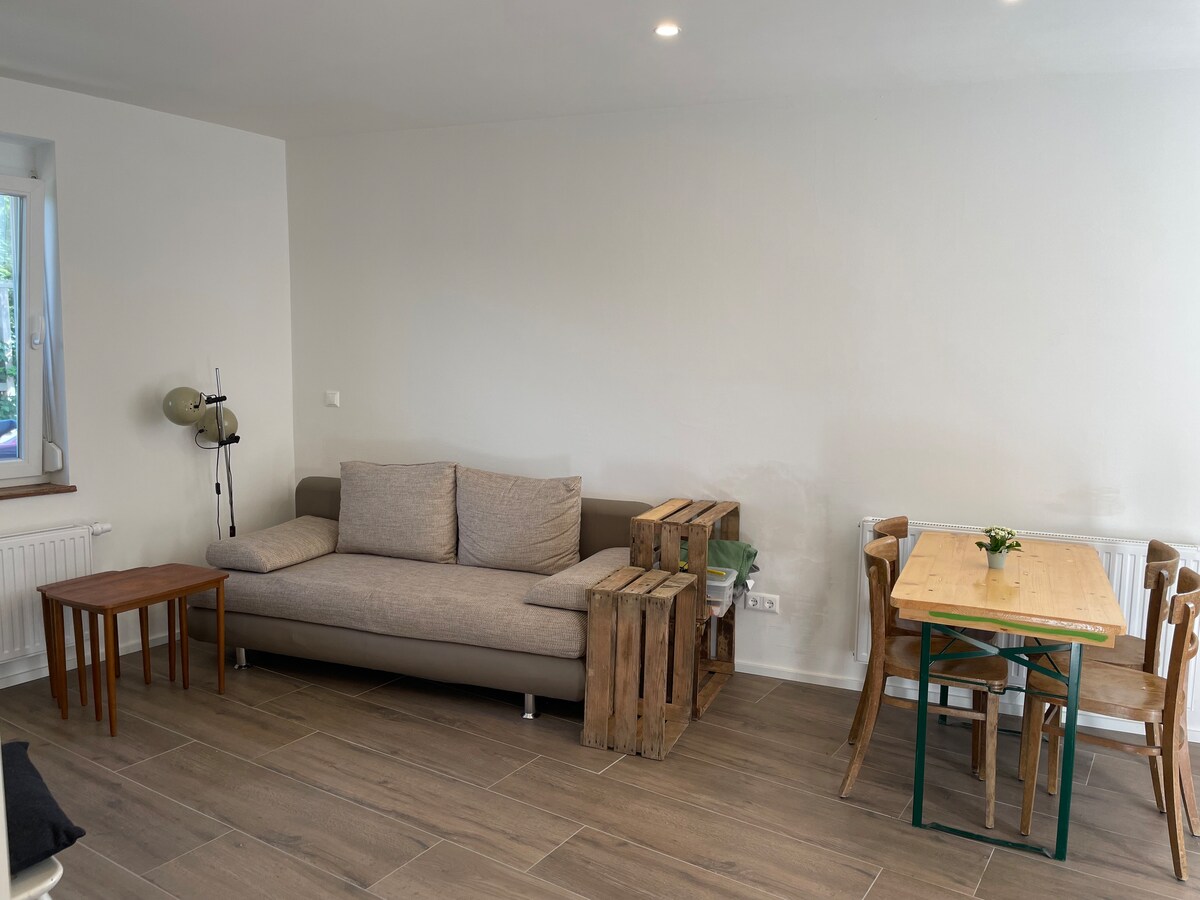 A modern living space is displayed, featuring a light grey couch positioned near a window. A wooden table with four chairs occupies one side, and two small wooden side tables are nearby. Natural light enhances the open atmosphere, complemented by warm-toned flooring.