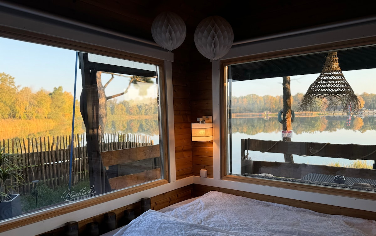 A tranquil scene is captured from the interior of the Tiny House, featuring expansive windows that frame a serene lakeside view. Soft natural light enters the space, illuminating a spacious bed with white linens. Decorative elements, such as a pendant light and plants, enhance the cozy ambiance.