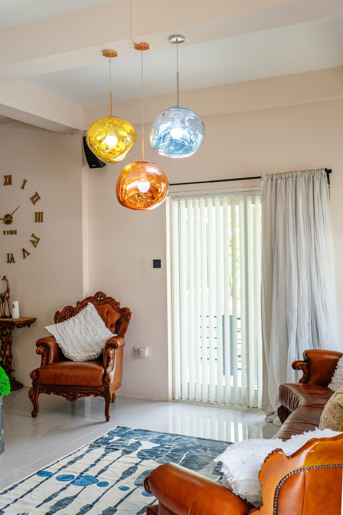 Colorful pendant lights are suspended from the ceiling above a cozy seating area. A leather armchair with decorative pillows sits beside a sliding door, which is partially covered by sheer white curtains that allow natural light to brighten the space.
