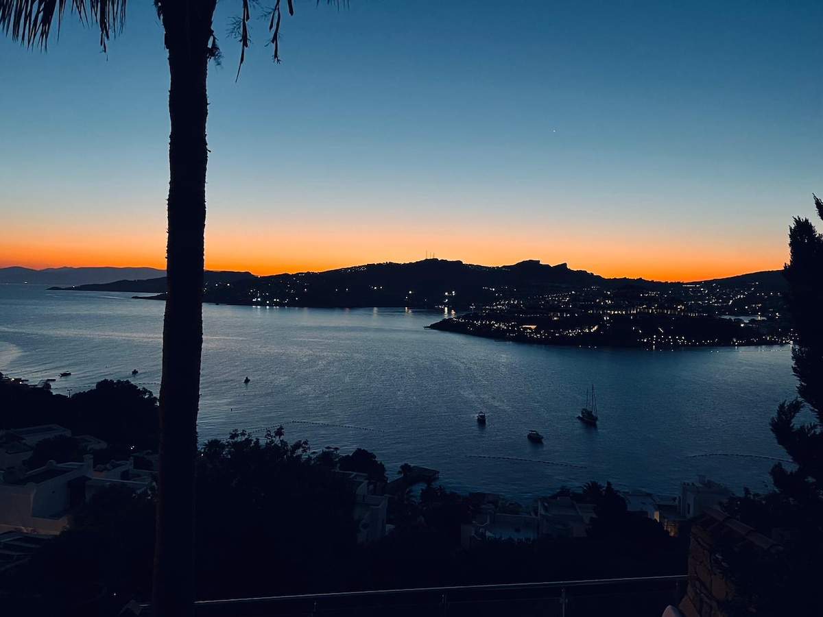 A breathtaking view captures the tranquility of twilight over Tilkicik Bay. The horizon is softly illuminated with vibrant oranges and blues, while silhouettes of hills and city lights reflect on the calm water. Boats float gently, adding to the serene coastal ambiance.