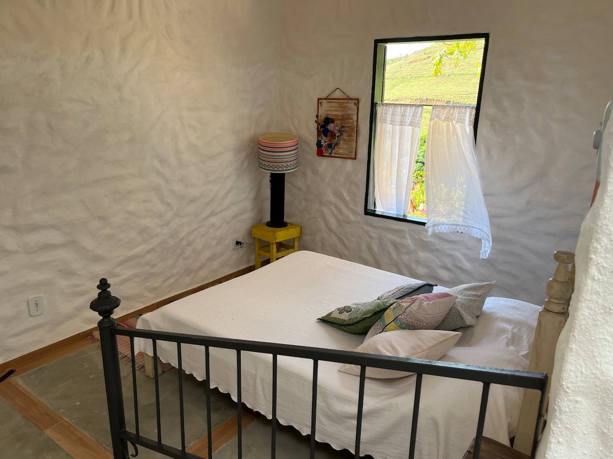 A rustic bedroom features a spacious bed dressed in white linens and accented with colorful pillows. A small lamp stands on a yellow nightstand beside the bed. Light filters through a window adorned with a sheer curtain, illuminating the textured walls.