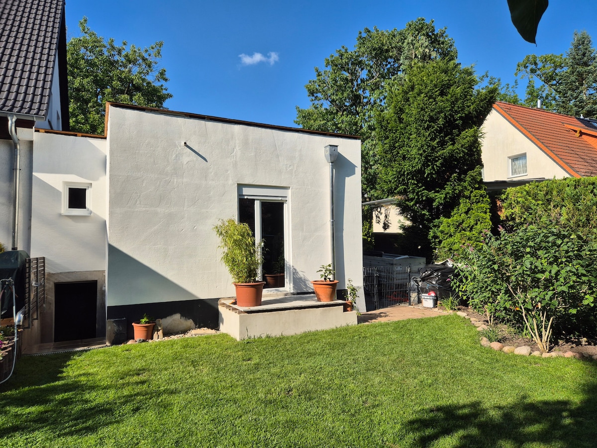 A private garden area is visible, featuring well-maintained green grass and potted plants near the entrance of the single-story rental unit. A cozy patio area is marked by a large window reflecting the surrounding greenery, with trees providing a natural backdrop.