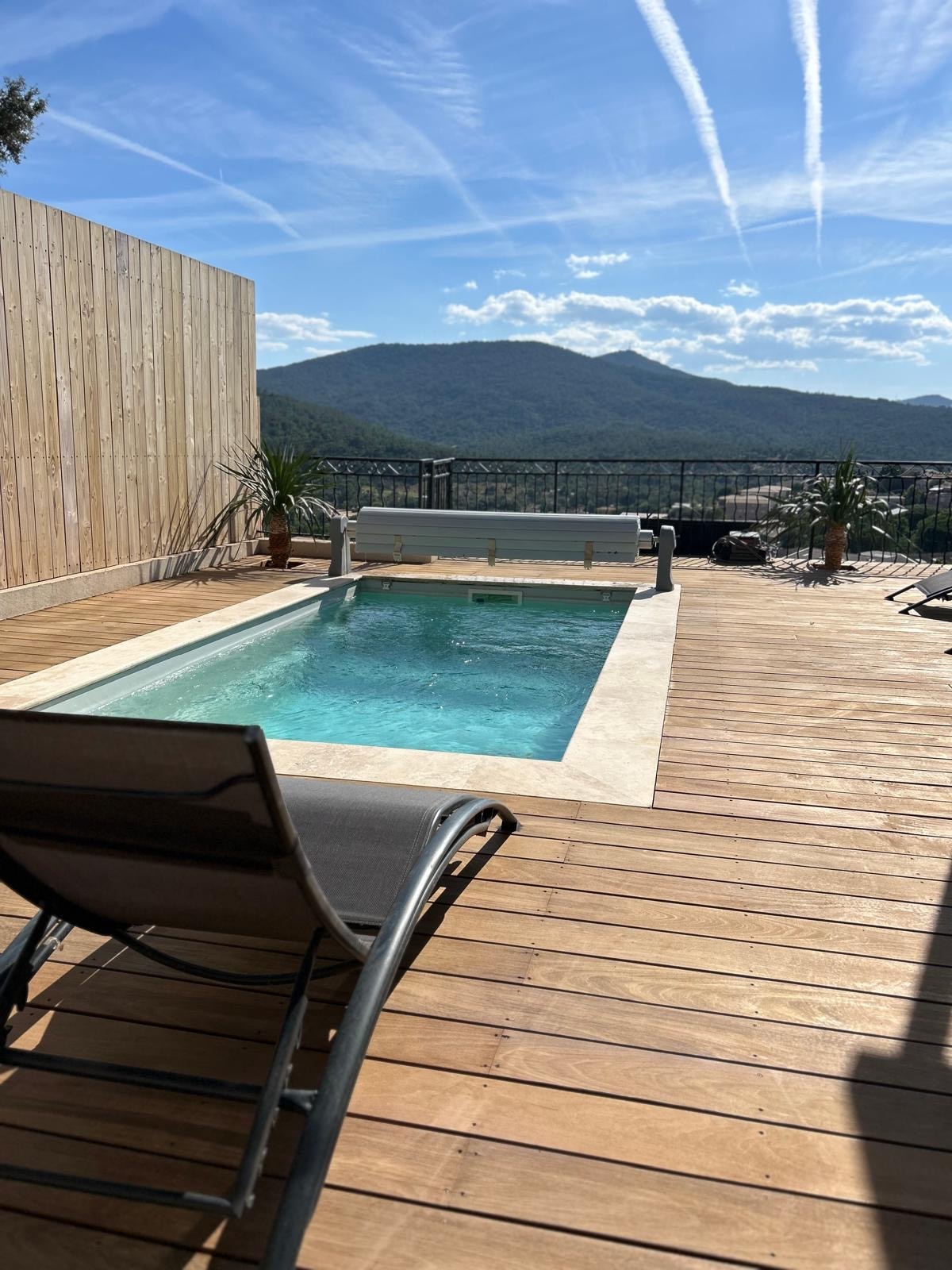 A serene pool area is surrounded by wooden decking, with a lounge chair positioned nearby. The clear water is visible against the backdrop of a distant mountain and a bright sky with wispy clouds. Potted plants add a touch of greenery to the space.