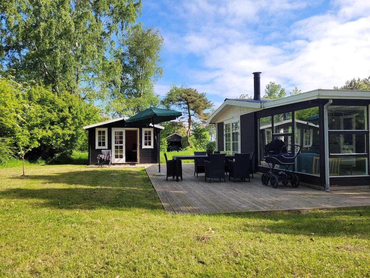 A spacious outdoor area is situated behind the house, featuring a wooden deck with seating arrangements. A black annexe with a canopy is visible, alongside greenery that frames the property. A stroller is parked on the deck, indicating family-friendly amenities.